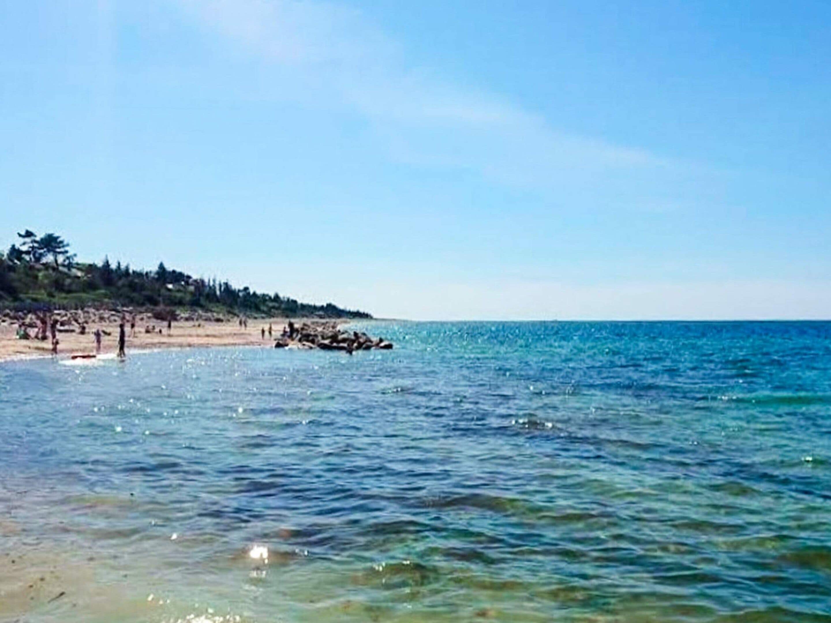Une plage à l’eau claire, un rivage sablonneux et une colline boisée en arrière-plan.
