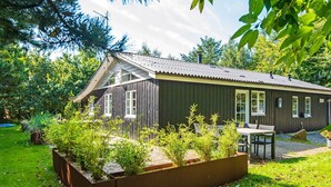 A wooden cabin with a sloped roof, a patio with seating, and a garden area with plants. - 4 Star Holiday Home in Ringkobing (Ringkobing)