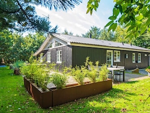 En stuga byggd av trä med ett sluttande tak, en uteplats med sittplatser och en trädgårdsdel med växter.