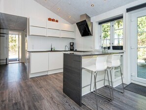 A modern kitchen with white cabinetry, a central island with bar stools, a flat-screen TV mounted above the island, and a large window providing natural light. - 4 Star Holiday Home in Ringkobing (Ringkobing)