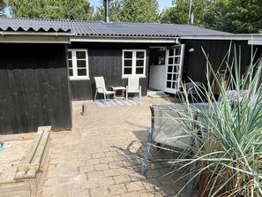 A wooden building with a metal roof, two white-framed windows, a patio with a table and chairs, and a large plant in the foreground. - 4 Star Holiday Home in Ringkobing (Ringkobing)