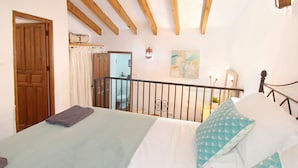 Quarto com cama, vigas de madeira, banheiro visível através de uma porta aberta e um quadro na parede.