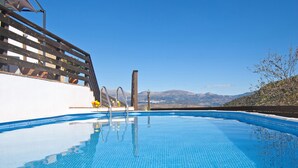 Piscina azul translúcida com um deck de madeira e vista para as montanhas ao fundo.