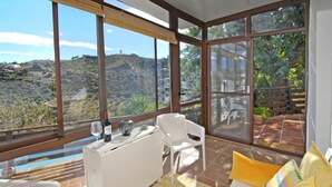 Quarto ensolarado com grandes janelas, um sofá branco, uma mesa com garrafa e copo, e vista para uma paisagem montanhosa.