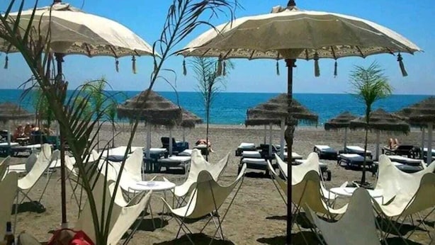 Uma praia com cadeiras de praia e guarda-sóis brancos, céu azul límpido e o oceano ao fundo.