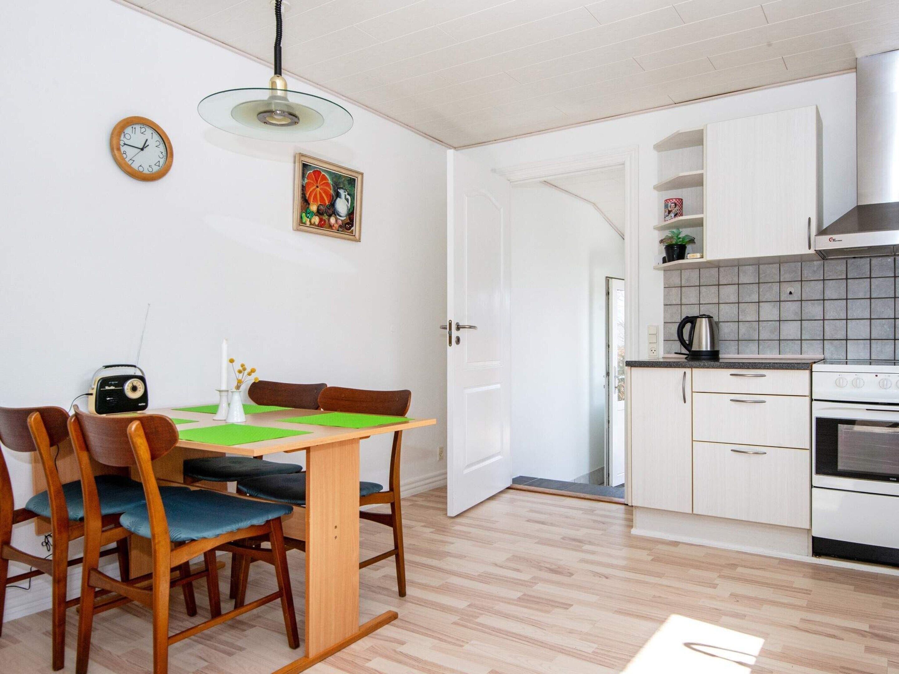 A compact kitchen with white cabinets, a stove, and a microwave. There's a dining table with chairs and a radio. A painting hangs on the wall, and a clock is visible.