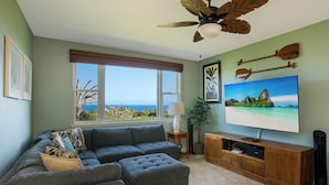 A living room with a sectional sofa, ottoman, and a flat-screen TV on a wooden stand. - 🌴 Ocean View Townhome | Free Late Checkout 🌴 (Kailua Kona)