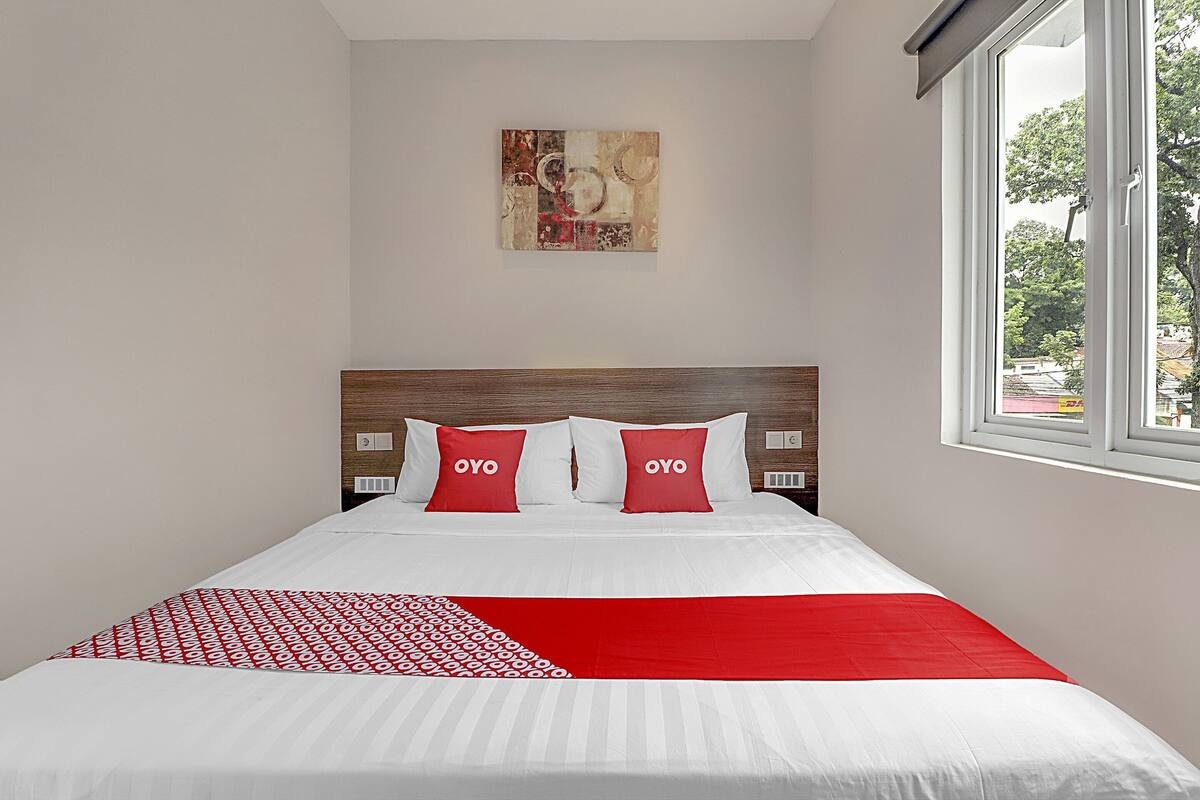 Un lit bien fait, avec une literie blanche et rouge, un plaid à motifs rouges et blancs, et un tableau accroché au mur.