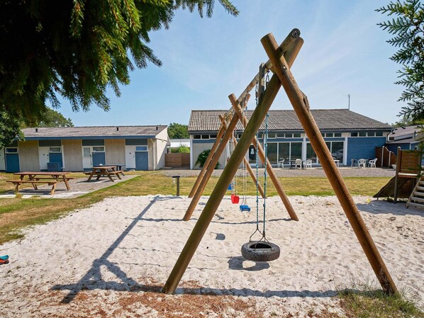 A playground with swings and a tire swing, picnic tables, and a building with a tiled roof in the background. - 4 Person Holiday Home in Nexo (Nexø)