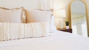Un lit bien fait, avec du linge de lit blanc et des coussins décoratifs, une lampe de chevet, une petite plante en pot et un grand miroir posé sur une table de chevet en bois.