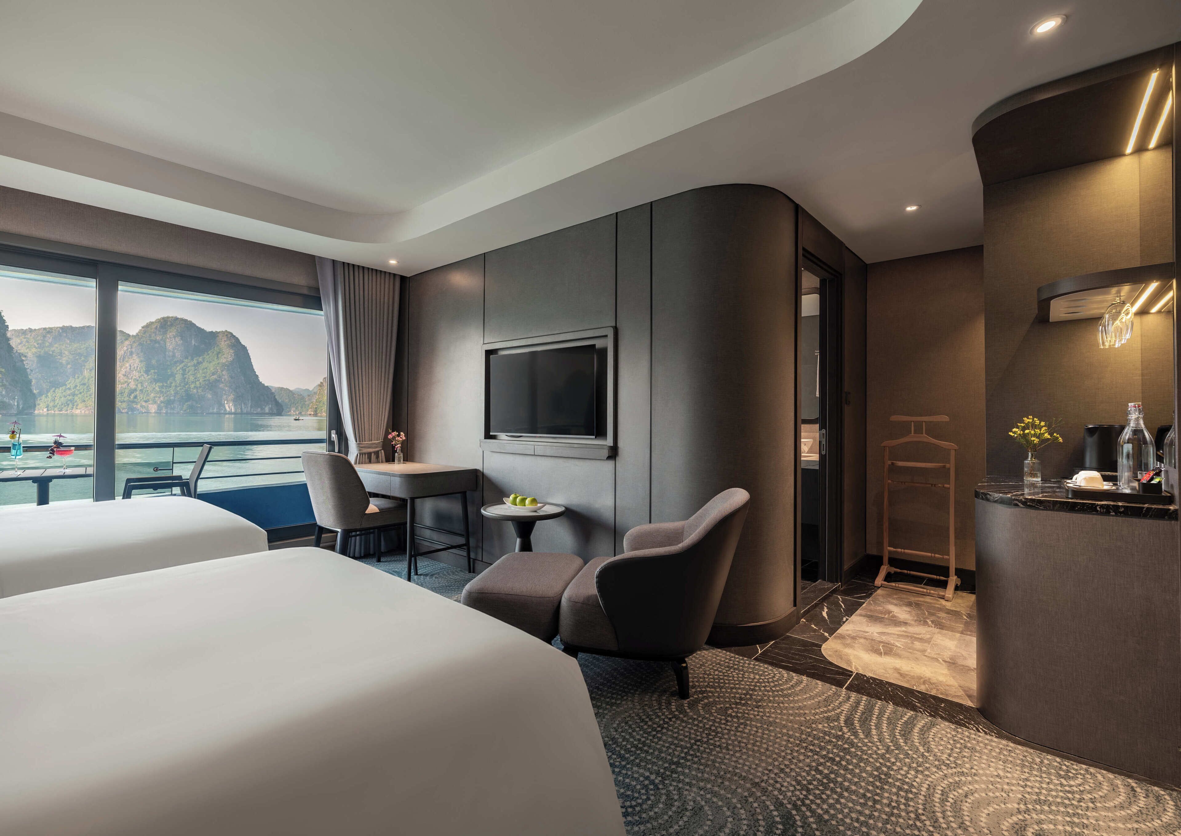 A modern hotel room with a large bed, a desk with a chair, a television, and a view of the sea.