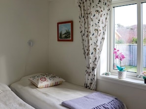 Un lit simple avec un coussin à motifs floraux, une fenêtre avec un rideau et une petite plante posée sur le rebord de la fenêtre.