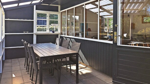 Un patio couvert avec une table et des chaises en bois, de grandes fenêtres et une vue sur l’extérieur.