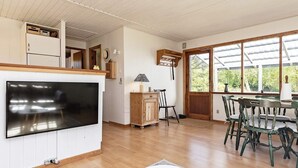 Un salon moderne doté d’un téléviseur à écran plat, de planchers en bois, d’un coin repas avec une table ronde et des chaises, et d’une grande fenêtre offrant une vue sur la verdure.