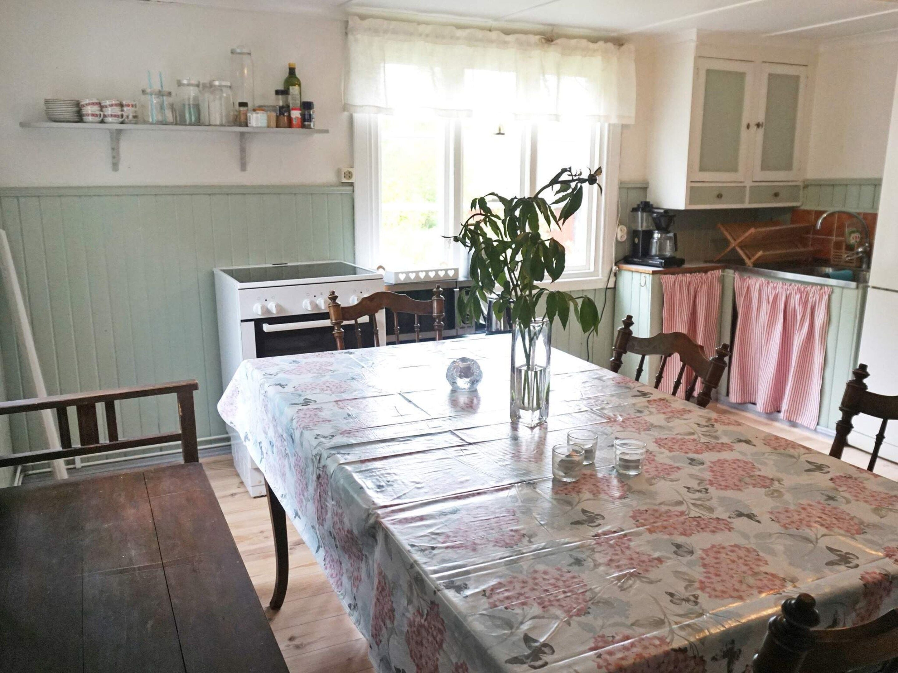 A dining area with a table set for two, a vase with a plant, and a kitchen in the background.