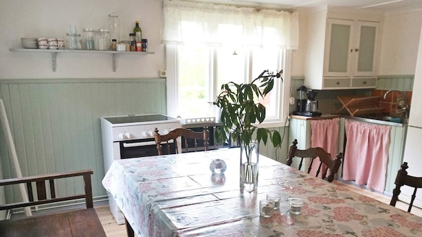 A dining area with a table set for two, a vase with a plant, and a kitchen in the background. - 6 Person Holiday Home in Hallabro (Hallaröd)