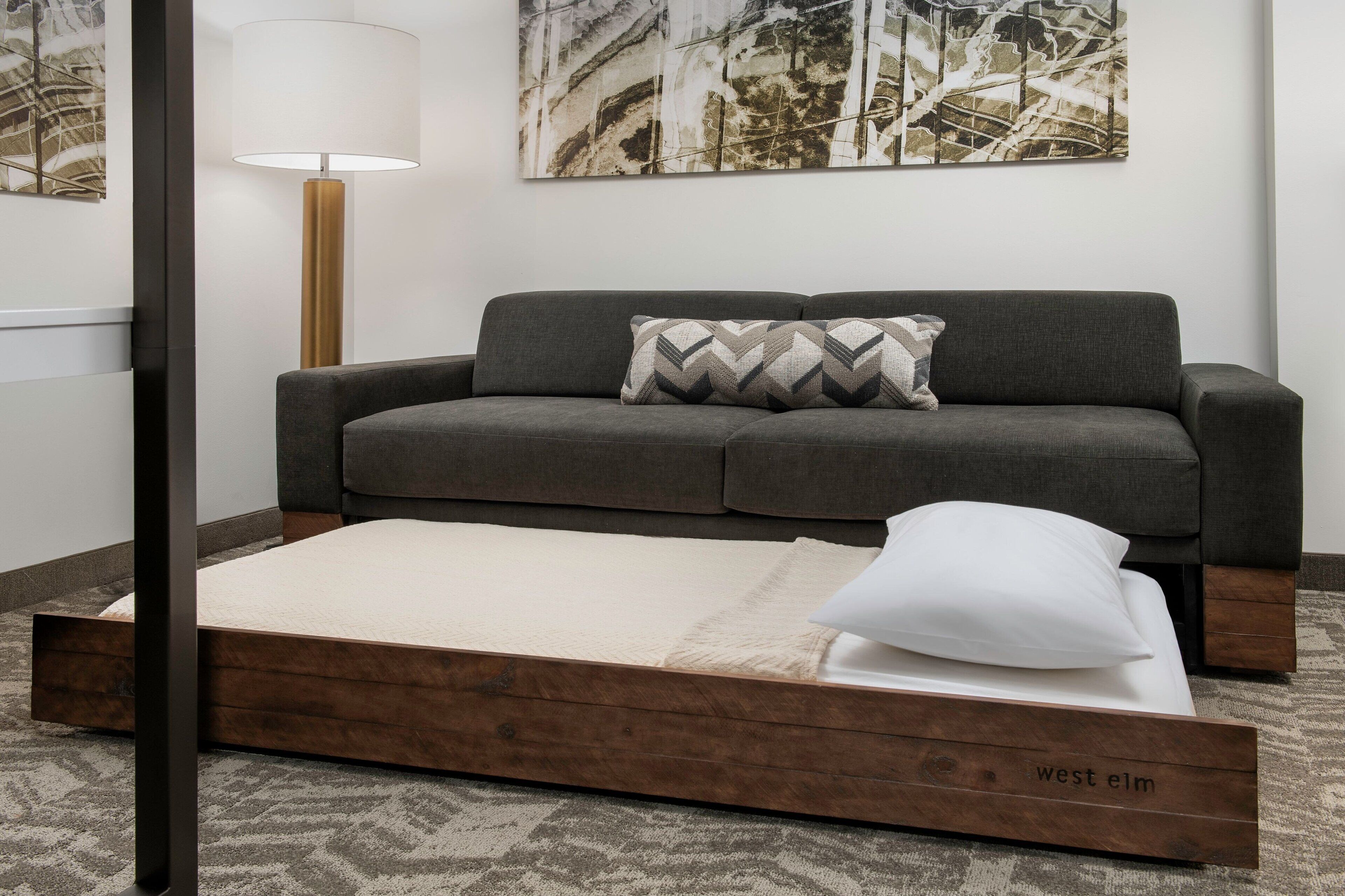 A modern room with a grey sofa, a wooden platform bed, a floor lamp, and abstract wall art.