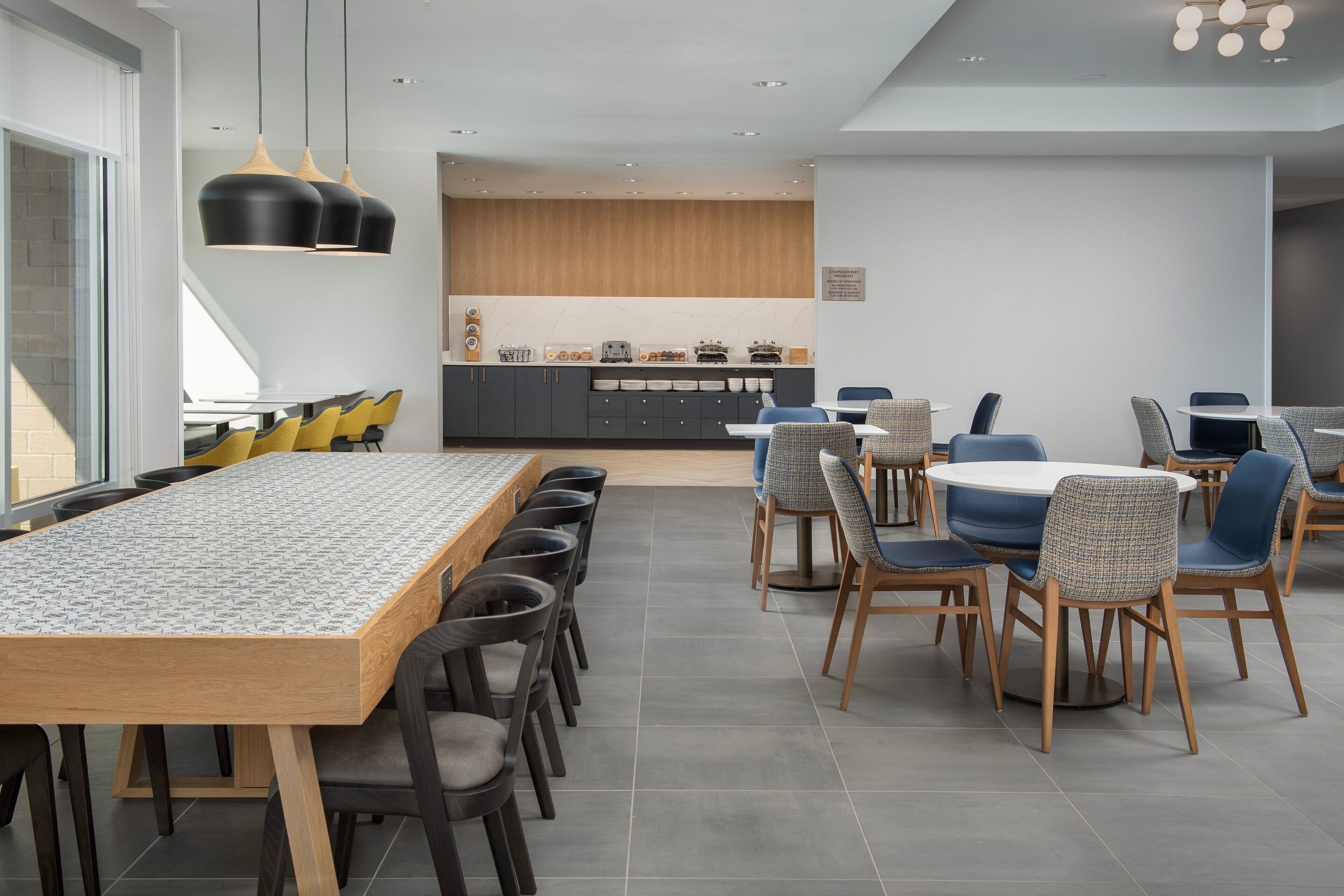 A modern dining area with a long wooden table and multiple chairs, a kitchen area with wooden cabinets, and pendant lighting.