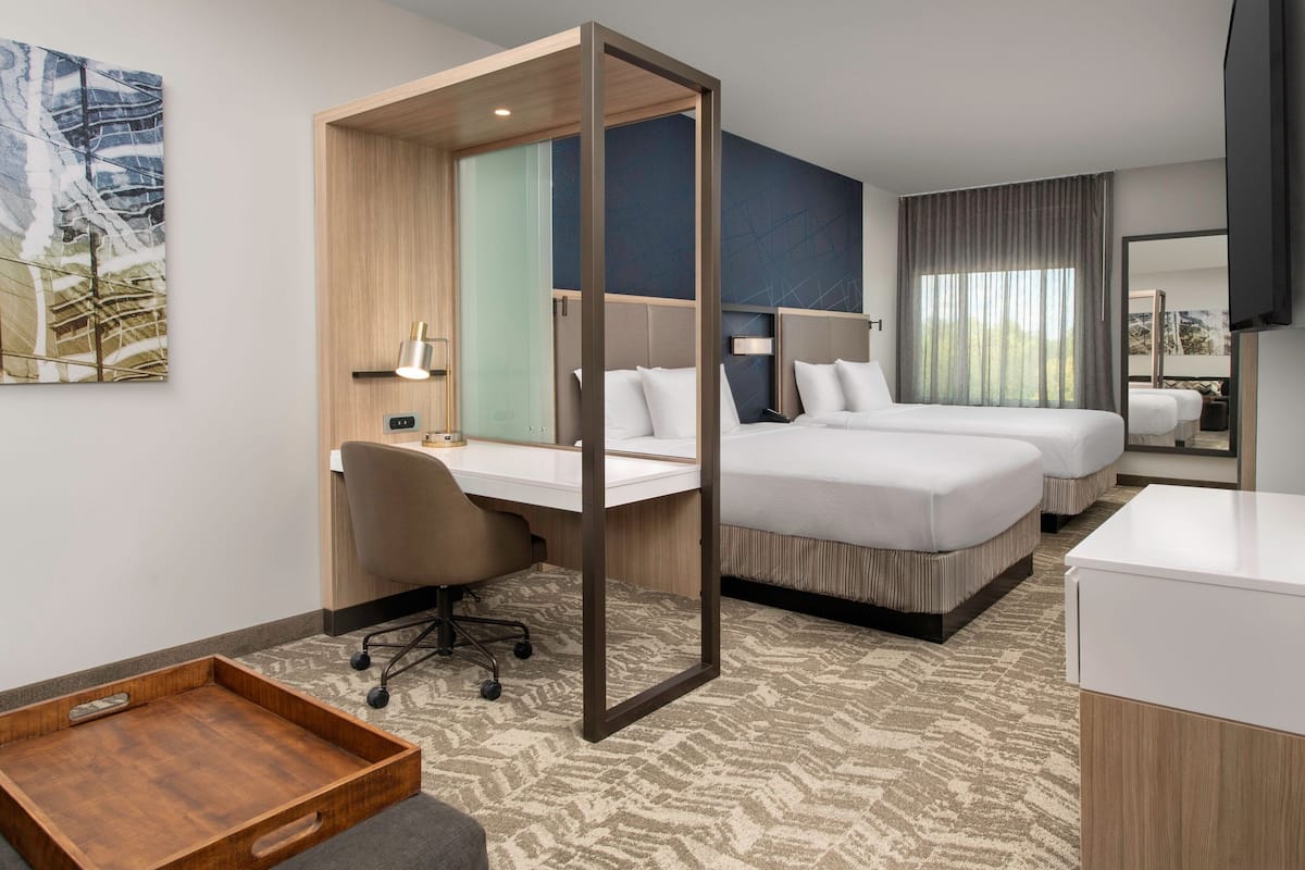 A hotel room with a desk, two beds, a wooden desk, a chair, a bedside table, a mirror, and a TV.