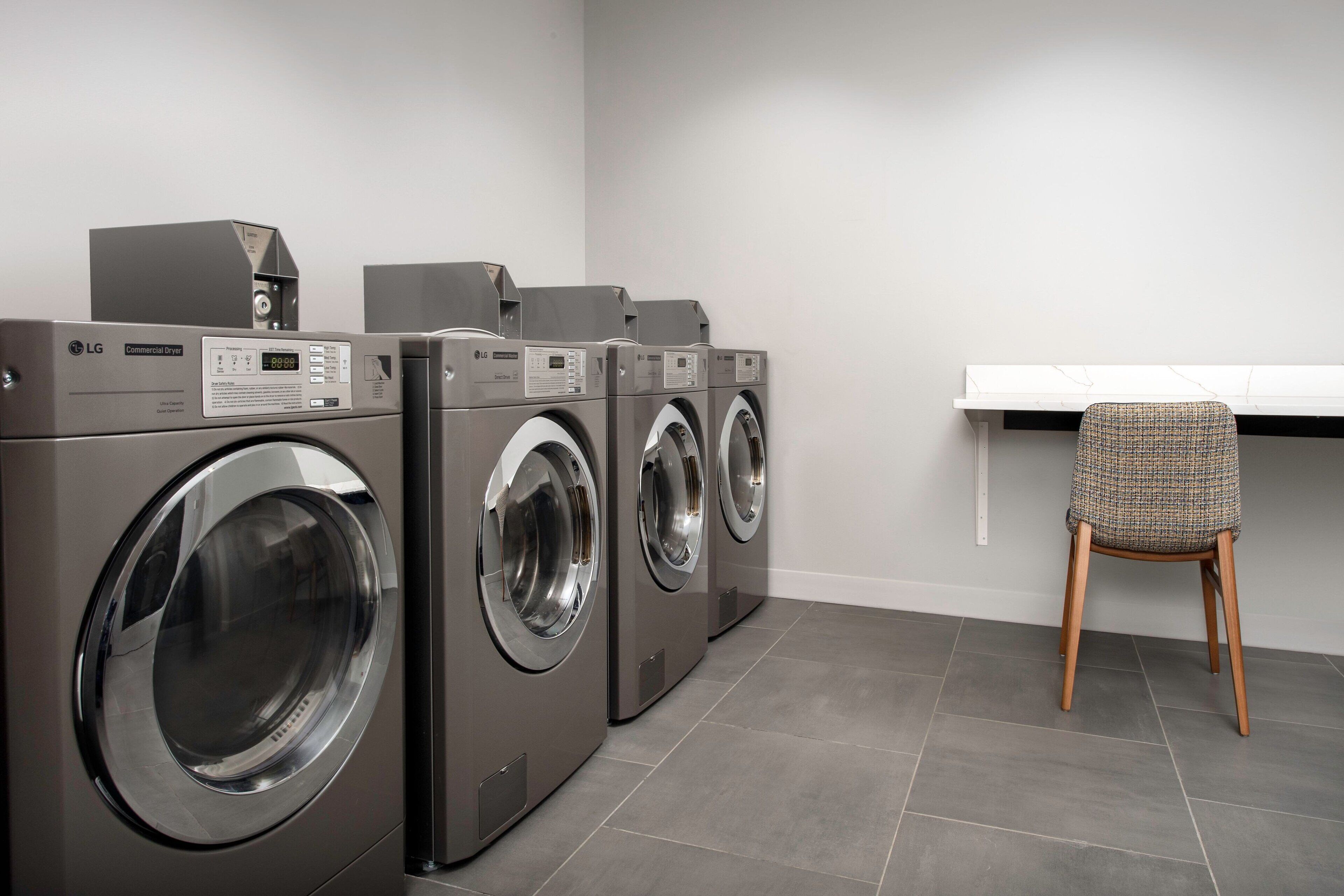 A laundry room with three LG commercial washers and dryers, a white shelf, and a wooden chair.