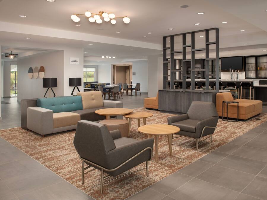 A modern living area with sofas, armchairs, and small tables, a kitchen area with bar seating, and a dining area with round tables.