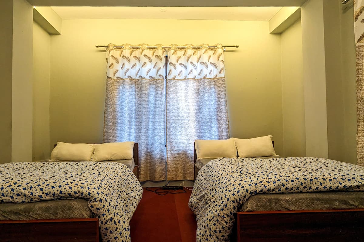 A small, single-room hotel room with two single beds, a window with patterned curtains, and a plain wall.