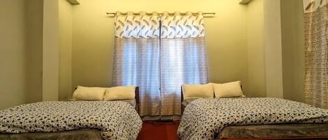 A small, single-room hotel room with two single beds, a window with patterned curtains, and a plain wall.