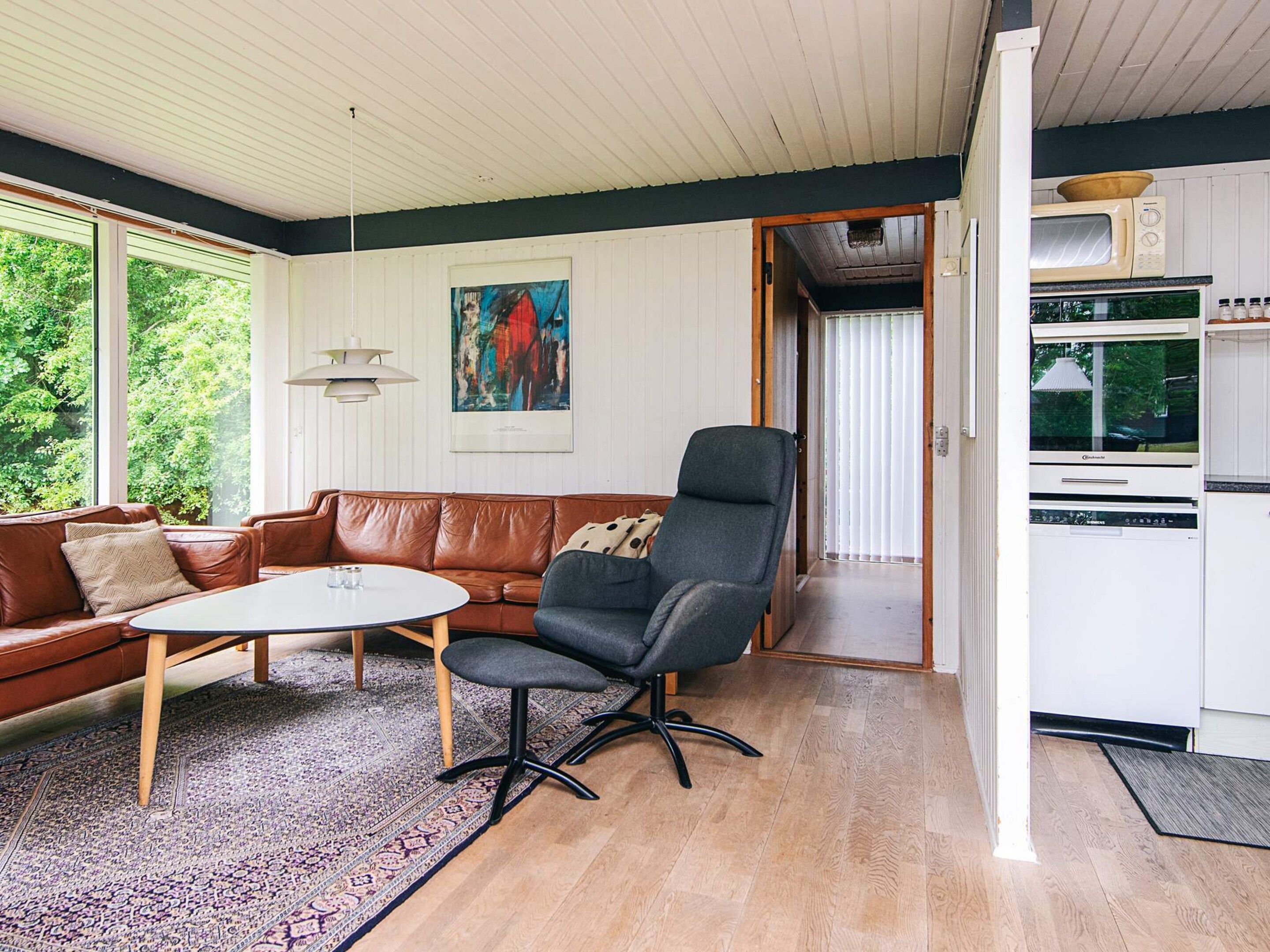 Una sala de estar moderna con un sofá de cuero marrón, un sillón gris y una mesa de centro blanca. En la pared, un cuadro abstracto de colores vivos y vistas al exterior a través de grandes ventanales.