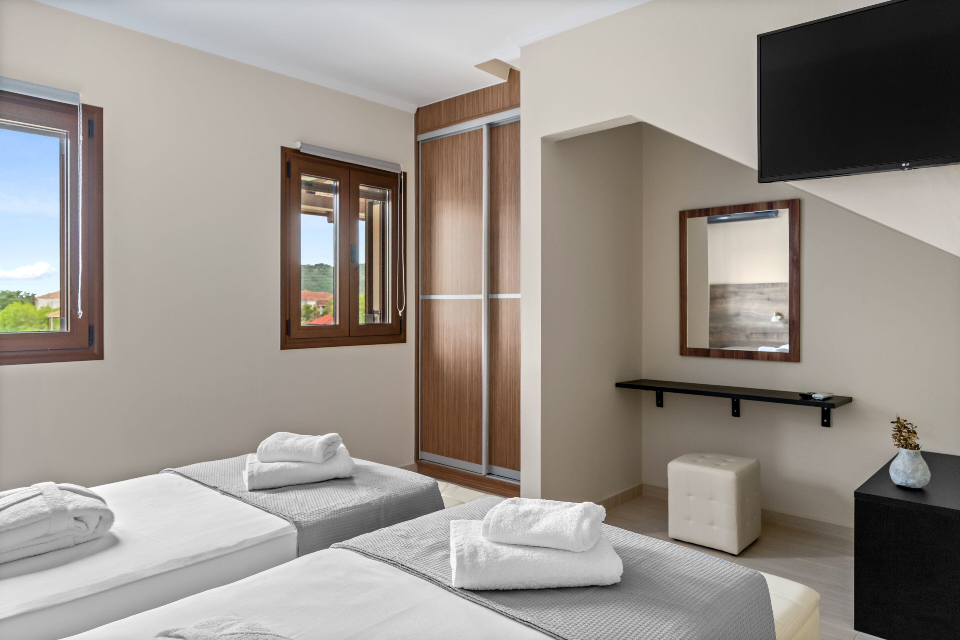 Quarto de hotel com duas camas, uma janela grande, uma porta deslizante, televisão montada e uma mesinha com um vaso.