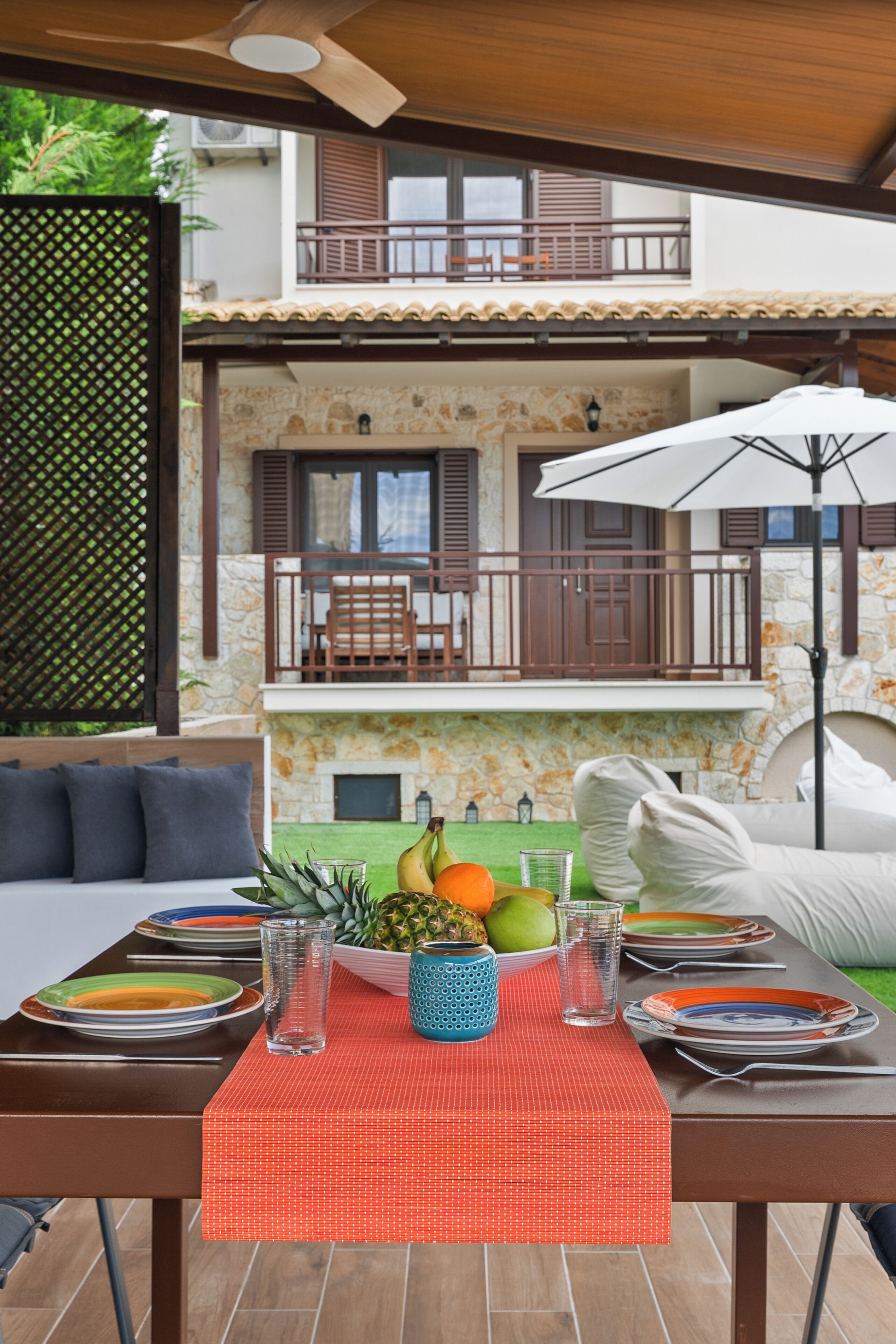 An outdoor dining area with a table set for two, a fruit bowl, and a view of a stone building with a balcony.