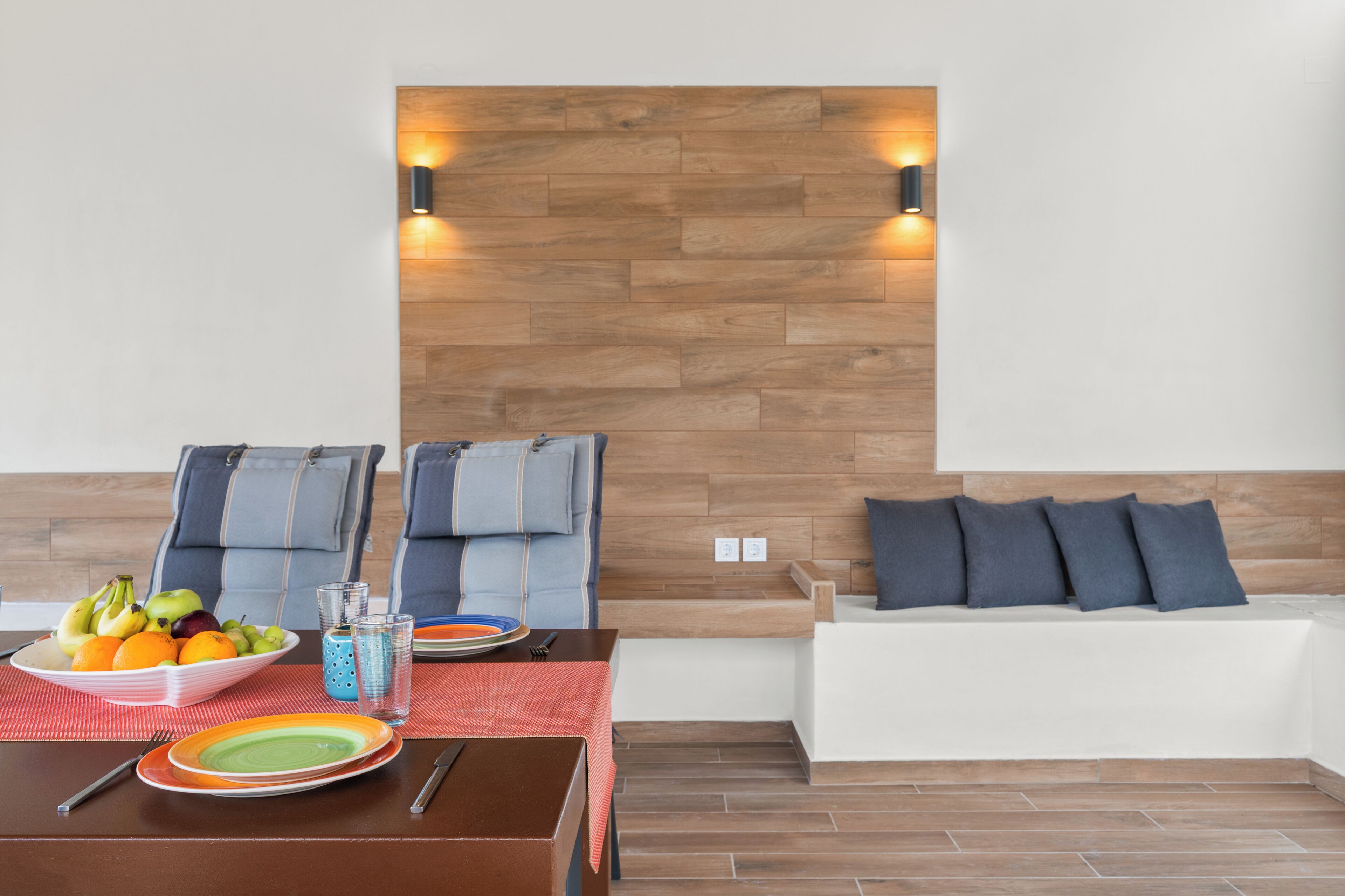 A modern room with a wooden accent wall, a sofa with cushions, a dining table with plates and fruit, and two wall-mounted lights.