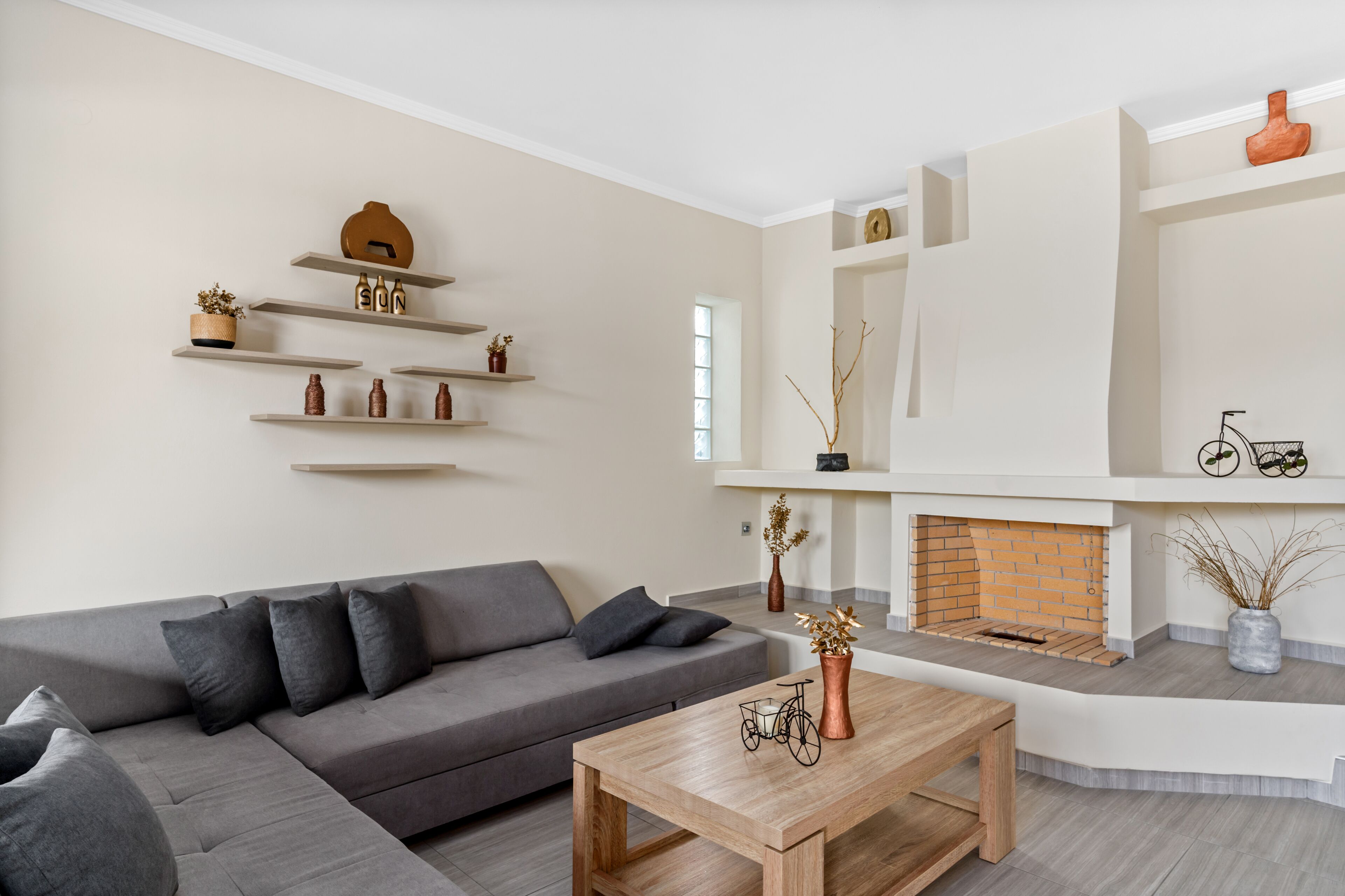 A modern living room with a grey sectional sofa, wooden coffee table, and a fireplace. Shelves with decorative items line the walls.