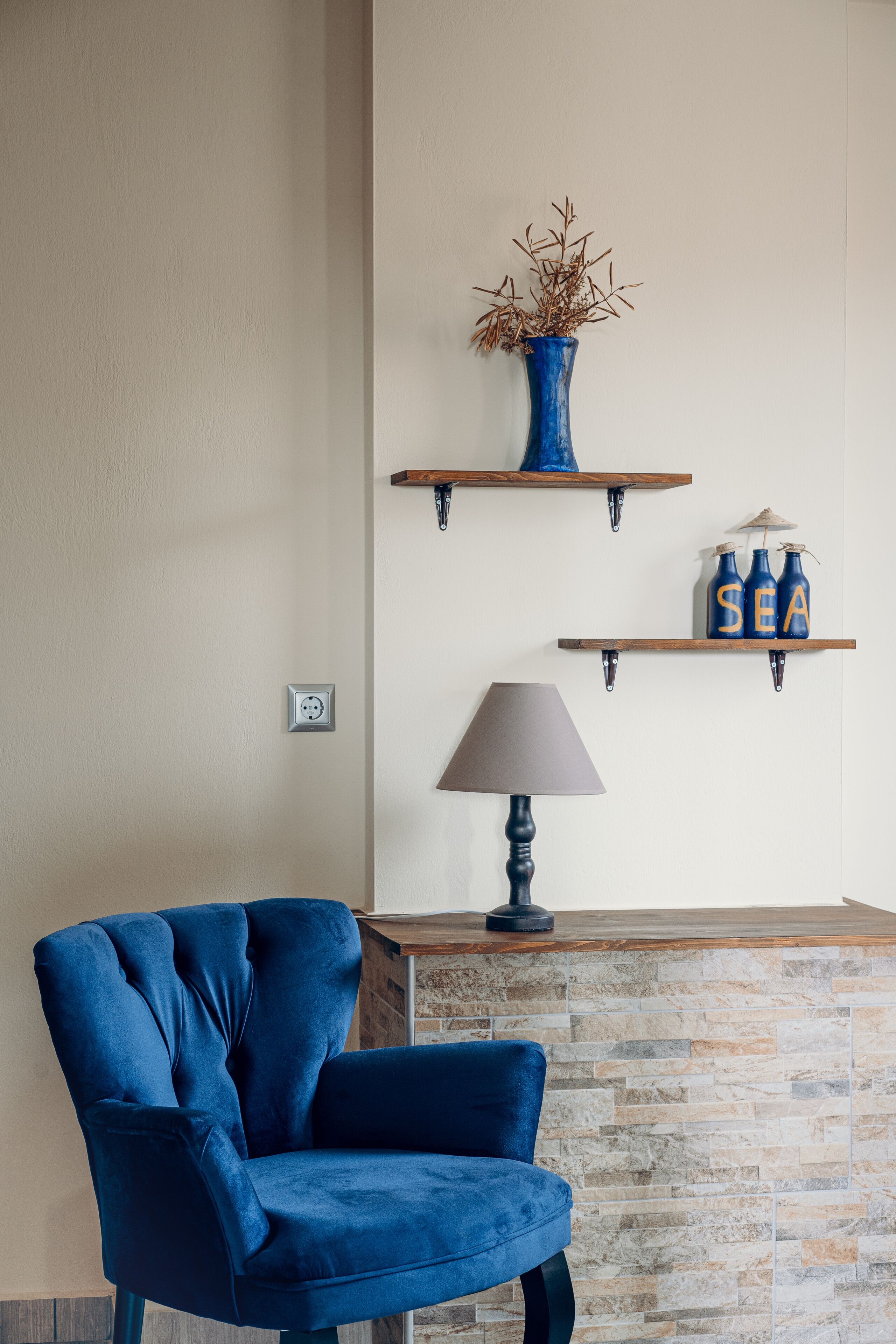 A room with a blue chair, a lamp, and two shelves with decorative items.