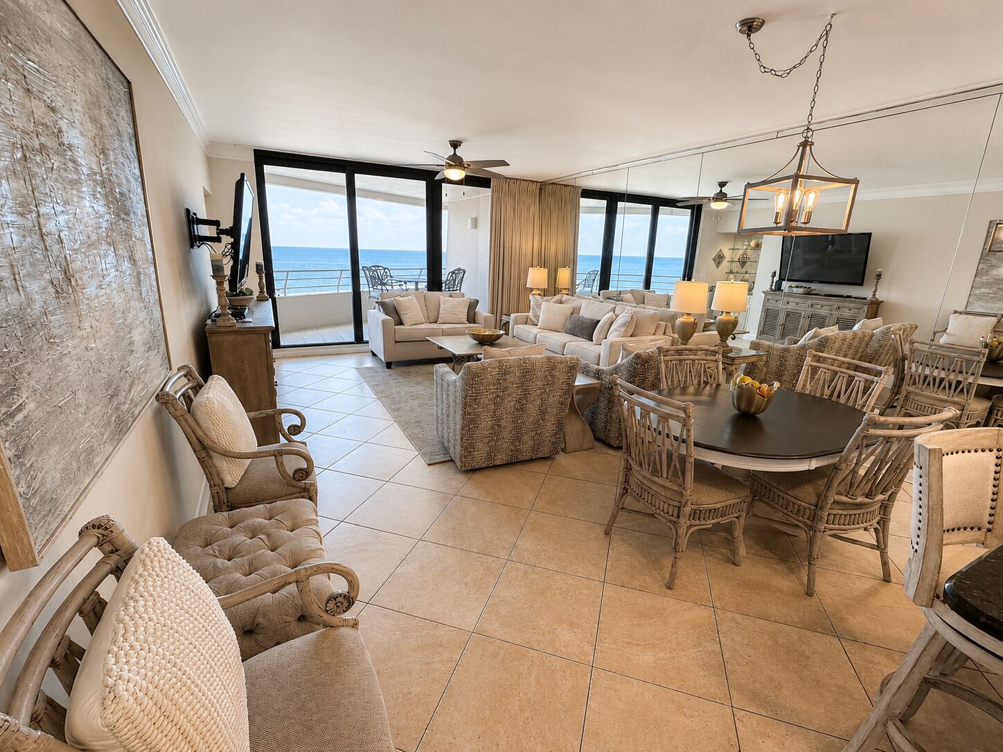 A spacious living area with a large window overlooking the sea, a dining table surrounded by wicker chairs, and a ceiling fan.