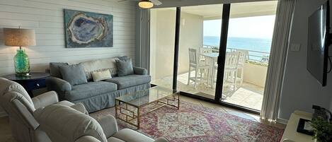 A living room with a sofa, armchair, and a coffee table. There is a painting on the wall, a lamp, and a view of the ocean through the sliding glass doors.