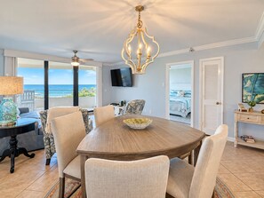 A dining area with a round table, chairs, a chandelier, a television, and a view of the ocean.