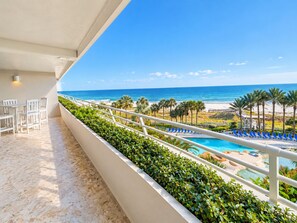 A balcony with a view of the ocean, a pool area with lounge chairs, and a dining area with chairs.