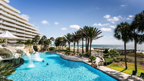 Una amplia piscina con cascada, rodeada de palmeras y vista a la playa.