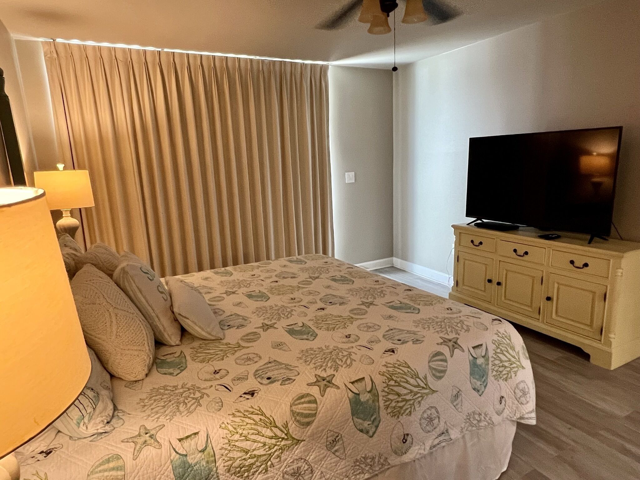 A bedroom with a bed, curtains, a television on a cabinet, and a ceiling fan.