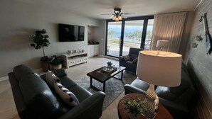 Una sala de estar con un sofá, una mesa de centro y un televisor. Se ve un ventanal con vistas al exterior.
