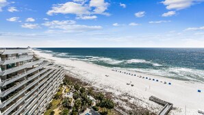 Un hôtel en bord de mer doté de nombreux balcons donnant sur l’océan, une plage de sable fin avec des chaises longues bleues, et un ciel dégagé.