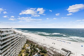 A beachfront hotel with multiple balconies overlooking the ocean, a sandy beach with blue lounge chairs, and a clear sky. - West 1102 2 Bedroom Gulf Front Condo with Stunning Views! (Pensacola)