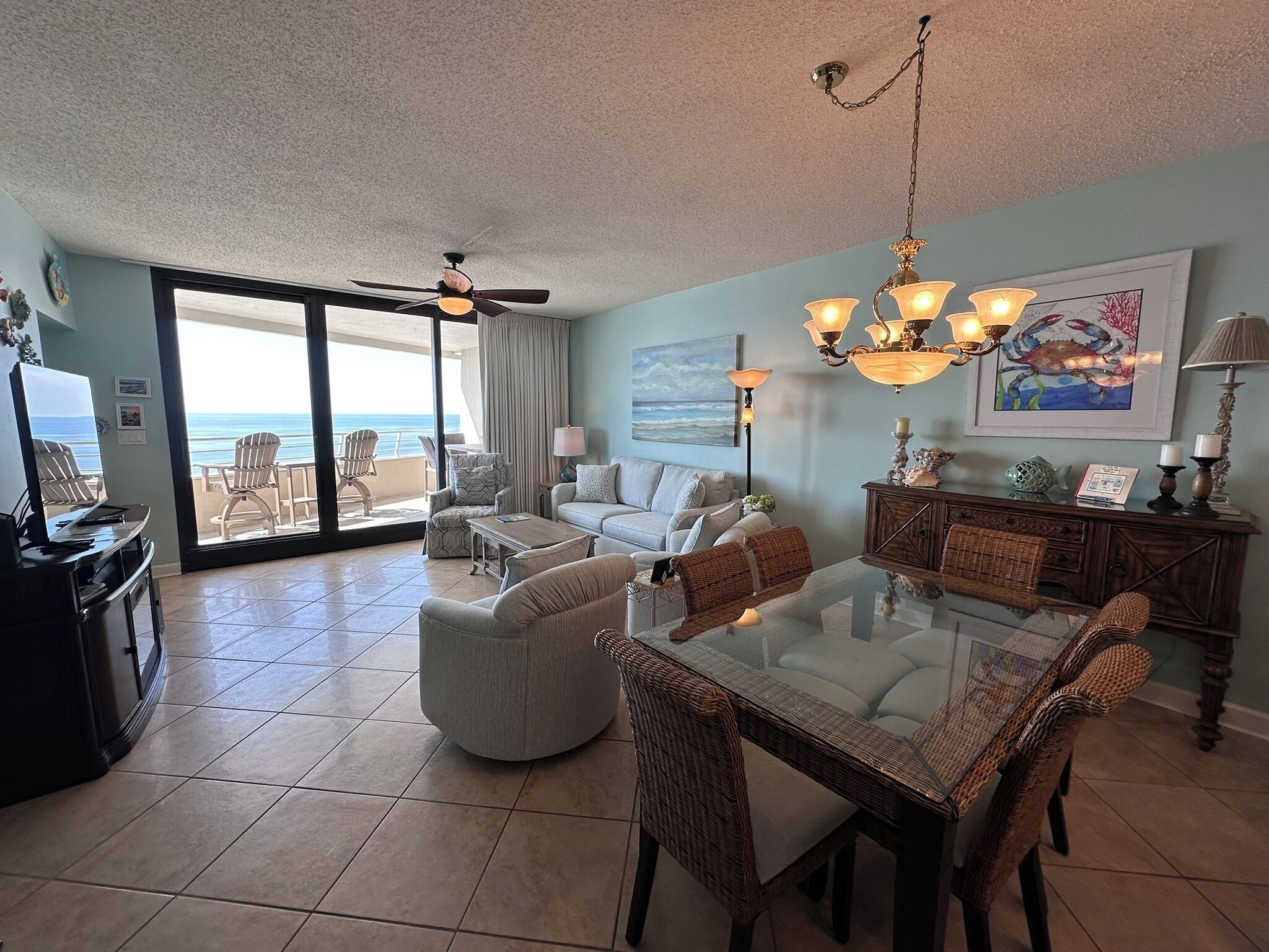 A spacious living area with a glass dining table, comfortable seating, a ceiling fan, and a view of the ocean through large sliding doors.