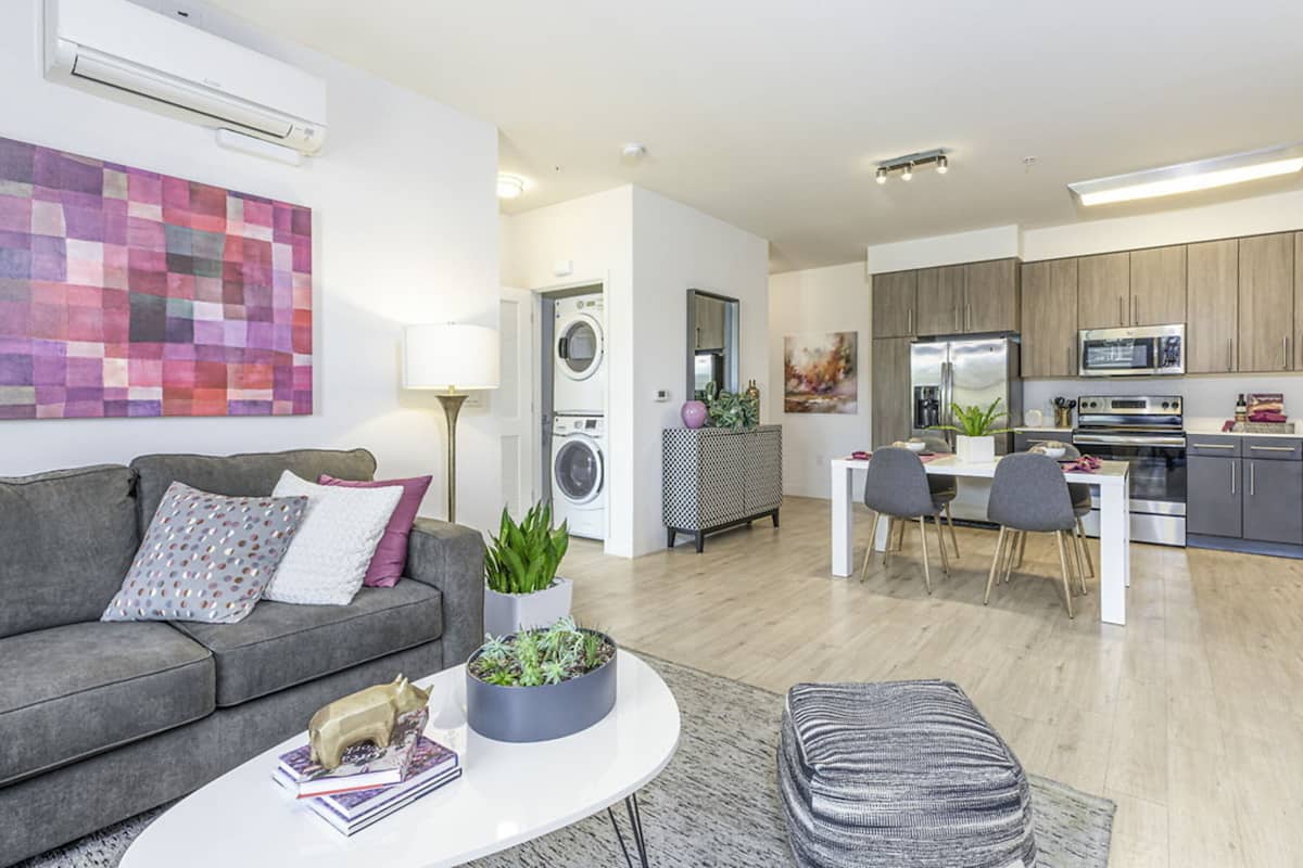 A modern apartment with a grey sofa, a round coffee table, a dining area, and a kitchen with grey cabinets and stainless steel appliances.