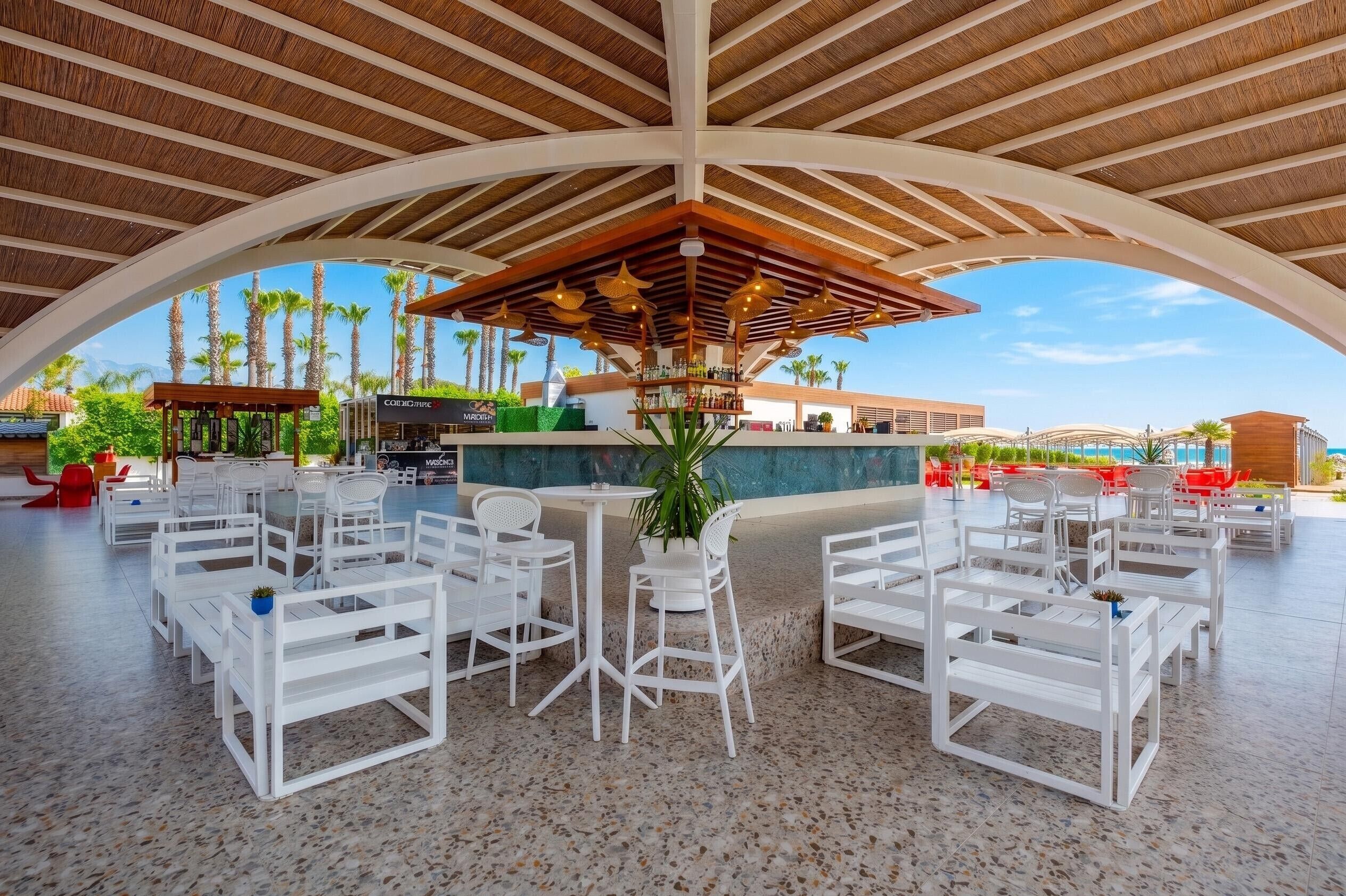 An outdoor dining area with white tables and chairs, a bar, and a view of palm trees and a pool.