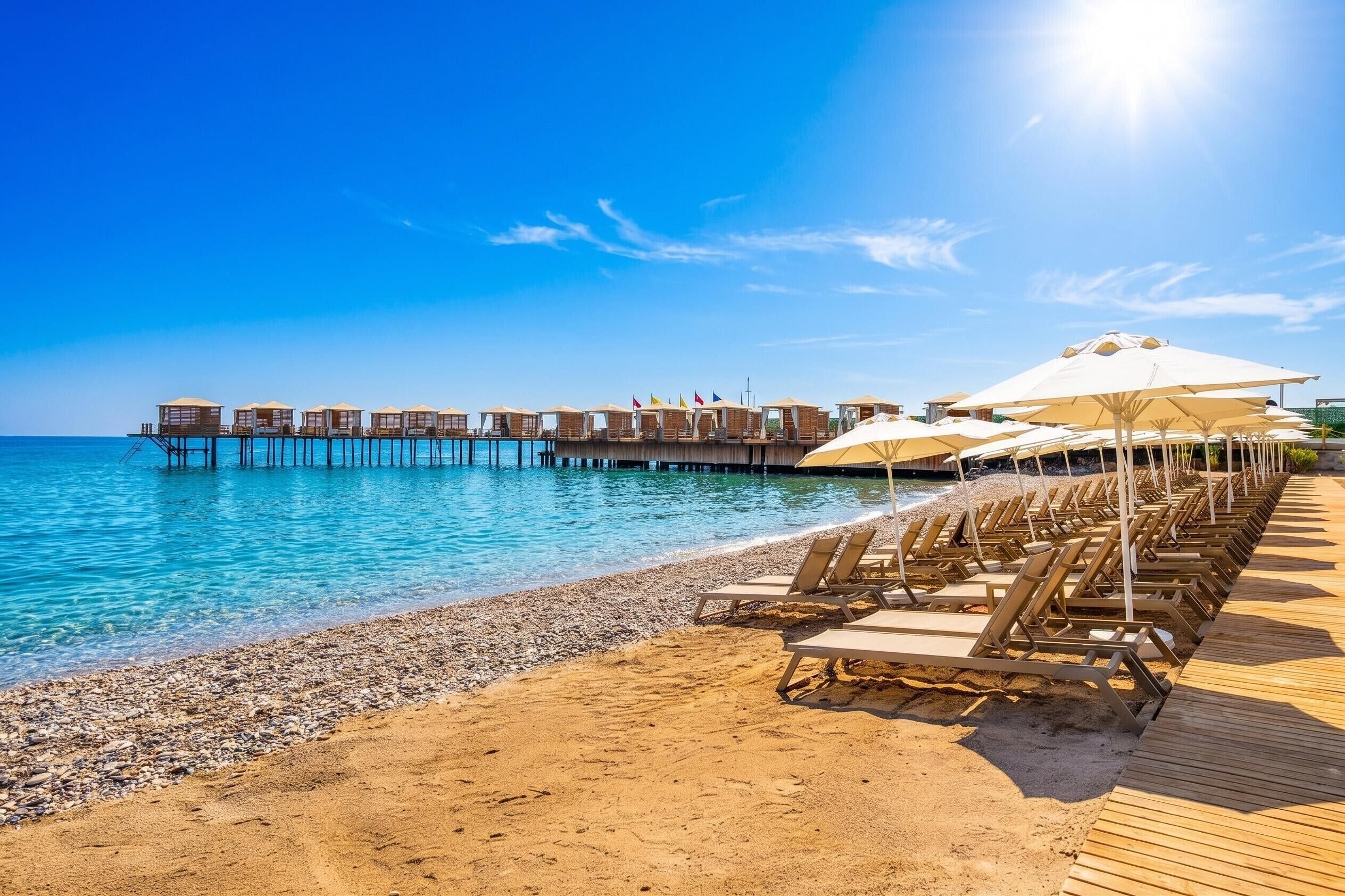 A beach with wooden deck chairs and umbrellas, a pier with overwater bungalows, and clear blue water.
