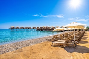 A beach with wooden deck chairs and umbrellas, a pier with overwater bungalows, and clear blue water. - Siu Collection Ultra All Inclusive (Kemer)