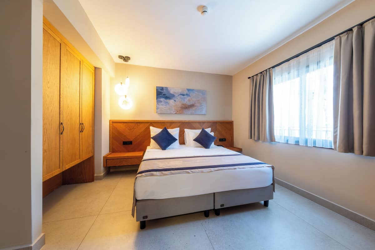 A hotel room with a bed, wooden wardrobe, wall-mounted light, and a painting.