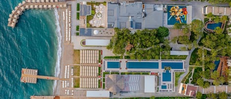 An aerial view of a resort with multiple swimming pools, a pier with sun loungers, and a large building with a rooftop pool area.