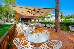 An outdoor dining area with white chairs and tables, a large umbrella, and a view of a building and trees. - Siu Collection Ultra All Inclusive (Kemer)